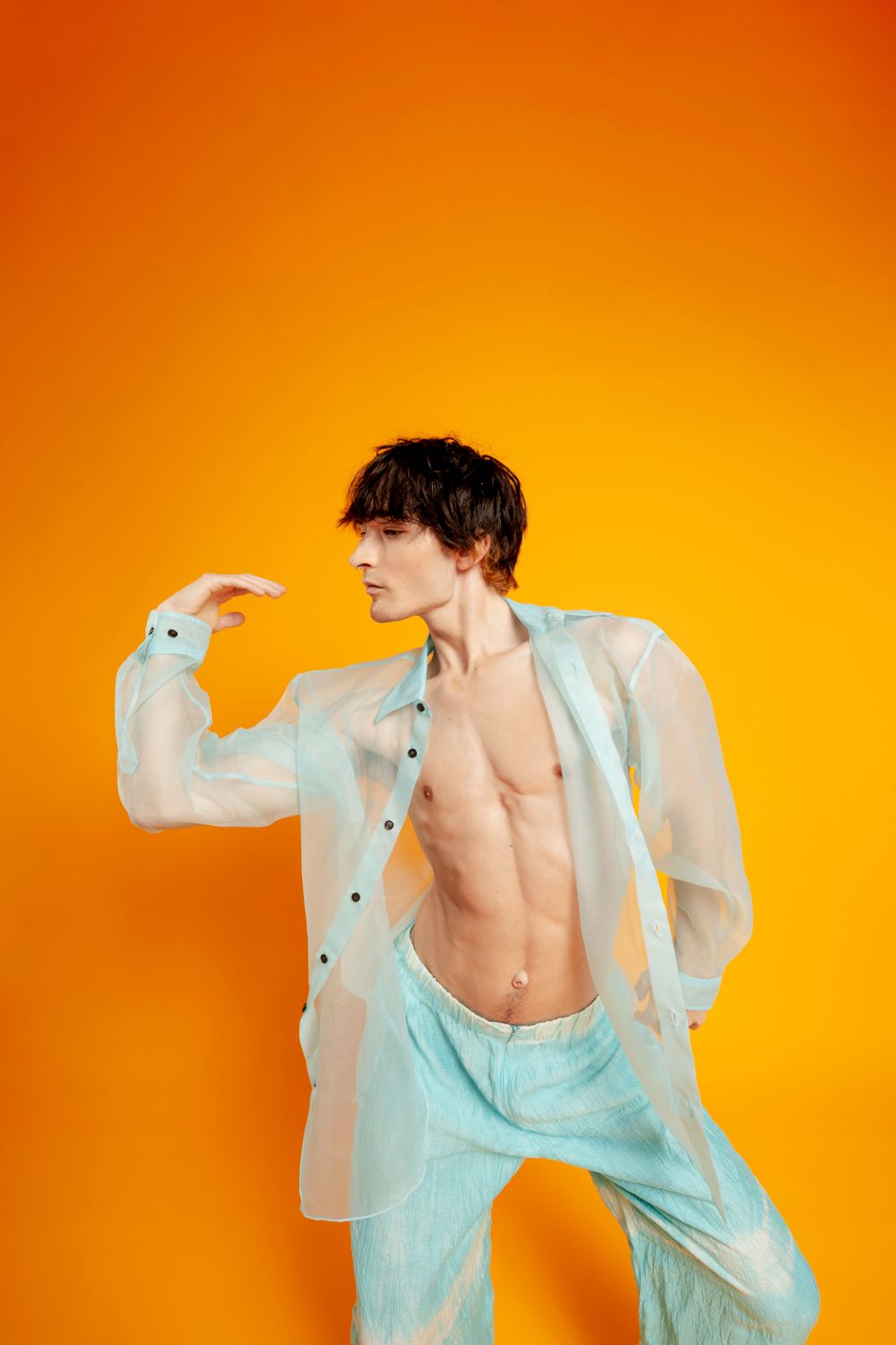Person in sheer white shirt and turquoise tank top poses dynamically against vibrant orange background.