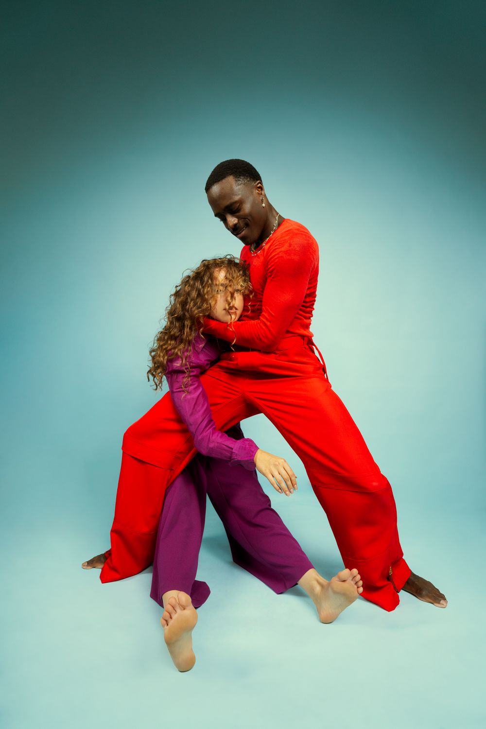 Two dancers in contrasting red and purple outfits perform a contemporary dance sequence against a teal backdrop.