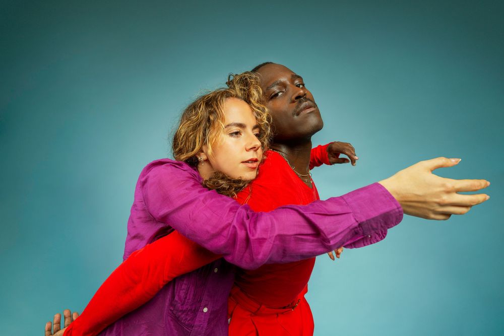 Two performers in vibrant purple and red outfits pose expressively against a teal background.