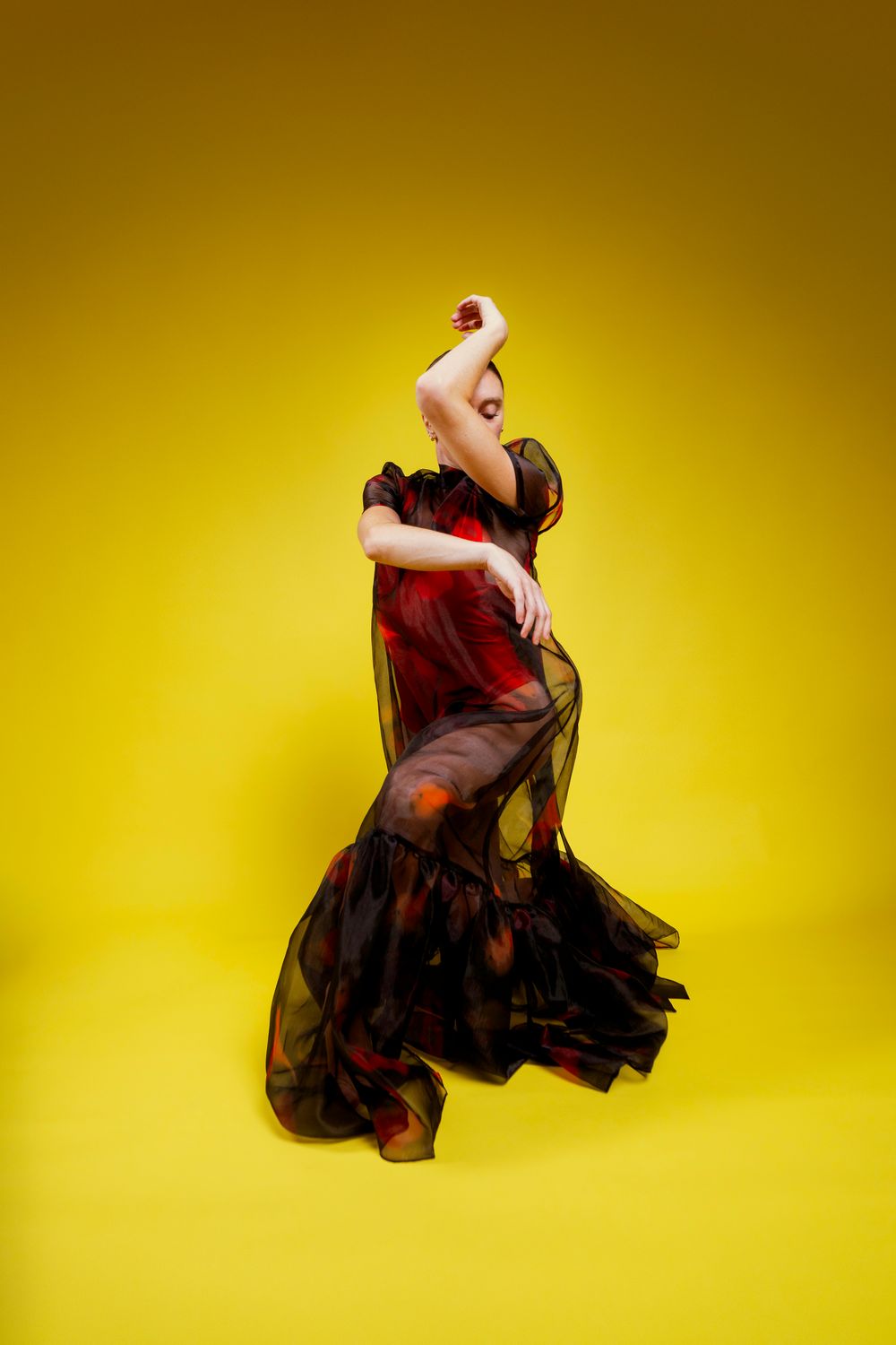 A dancer in a flowing black dress performs an expressive movement against a vibrant yellow background.