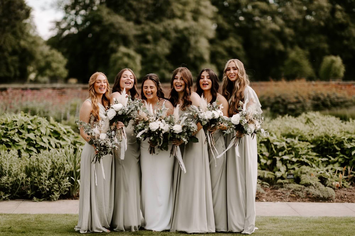A group in light gray dresses holds white and green floral bouquets while standing together outdoors.