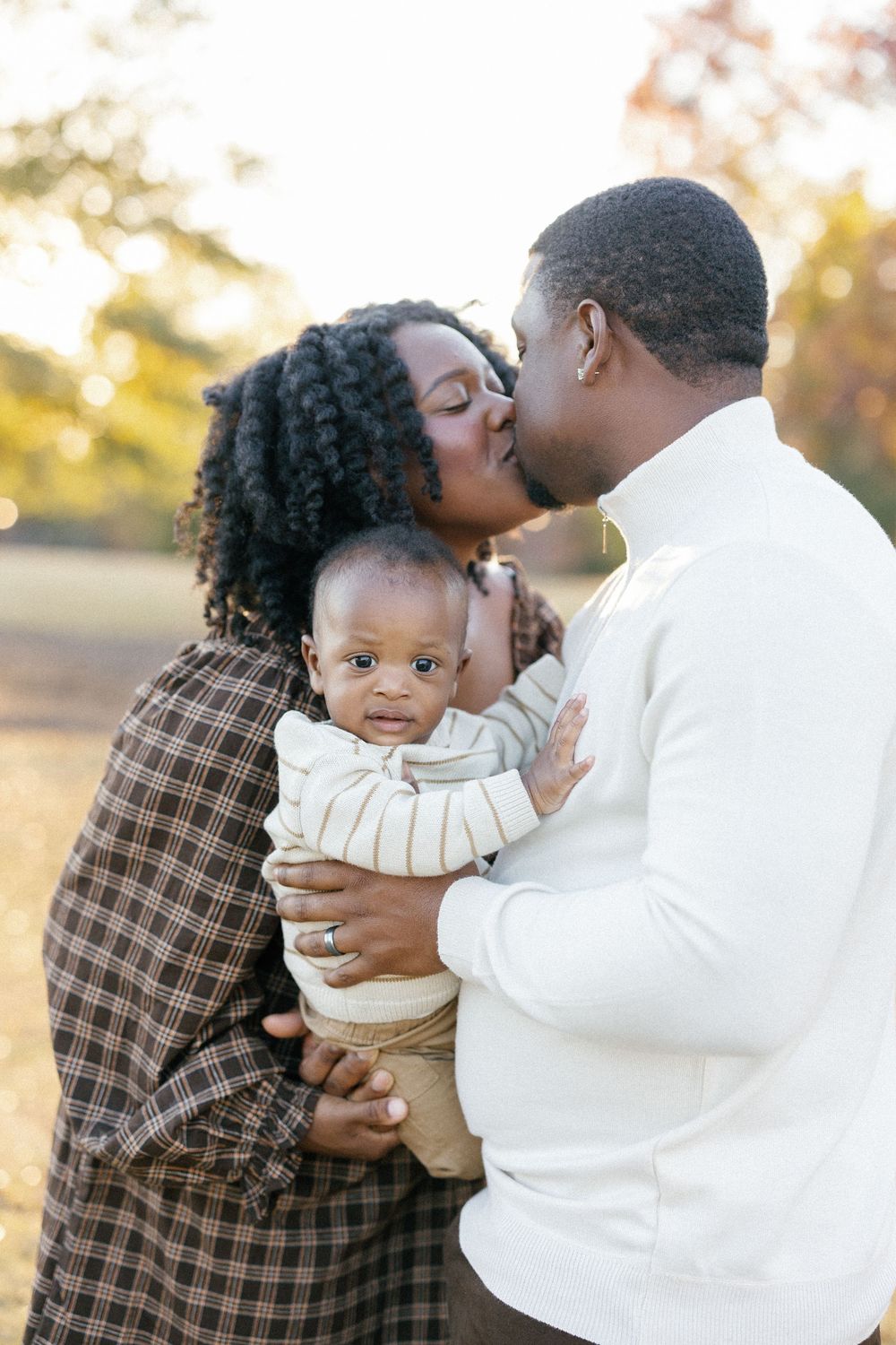 A tender family moment captured outdoors during golden hour with a baby in a cream-colored outfit.