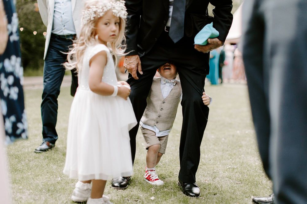 Wedding guests gather on a grassy lawn during an outdoor reception with children in white dresses playing.