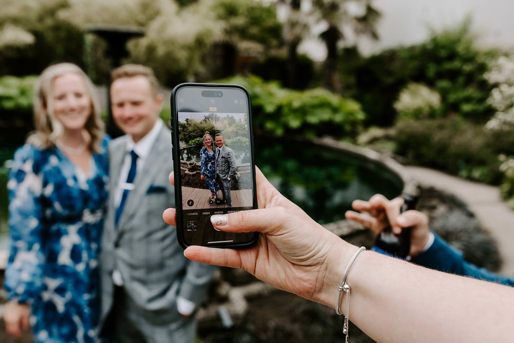 Hand holds smartphone to capture photo of couple in garden setting during outdoor photography session.