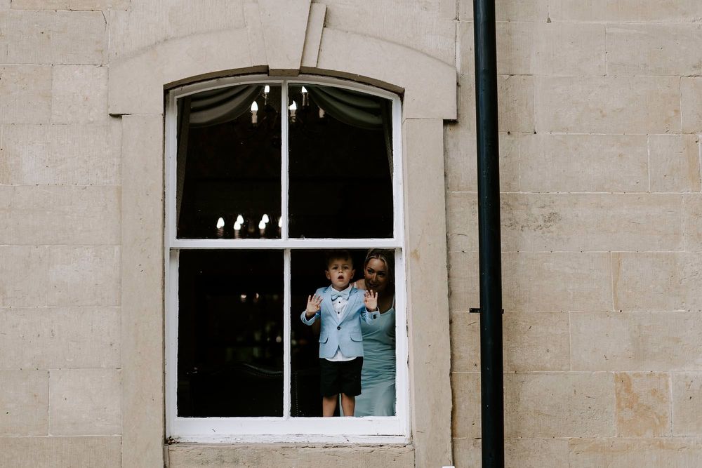 A child looks out through a large arched window in a stone building, with candles visible on the windowsill.
