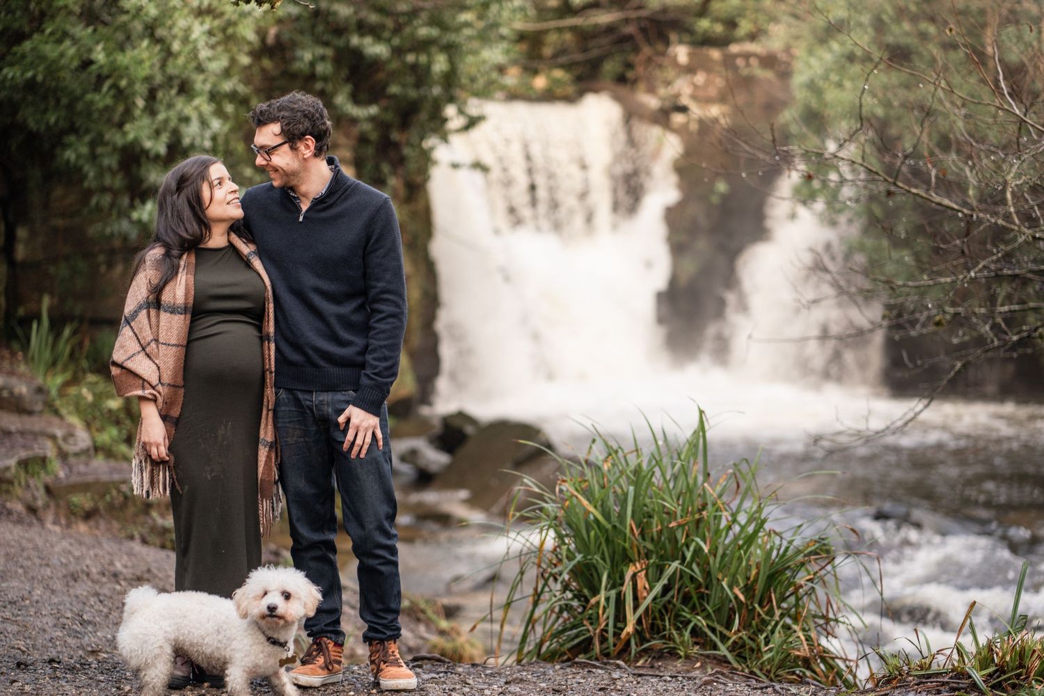 A couple and their small white dog stand together near a scenic waterfall surrounded by lush greenery.