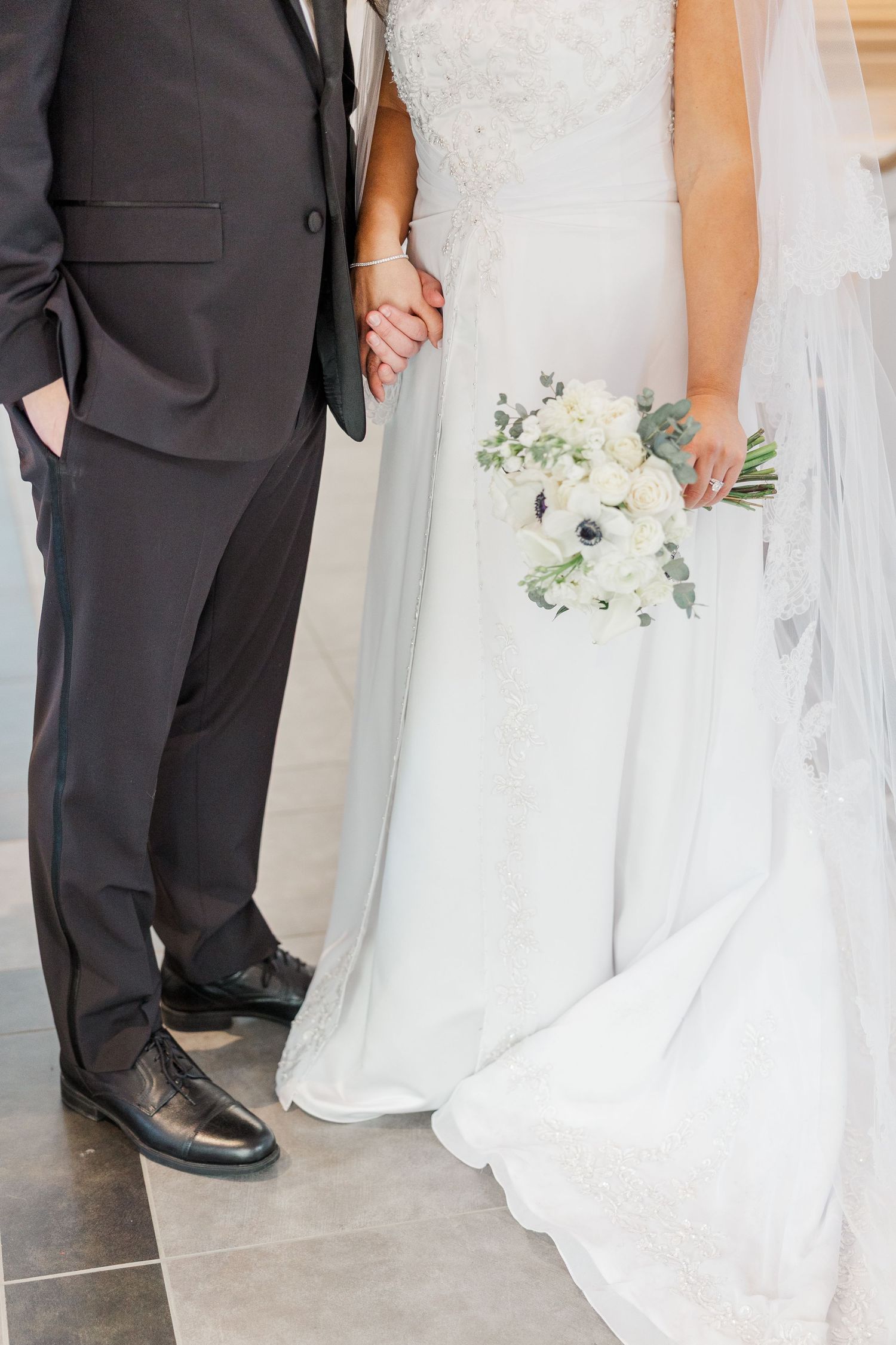 A wedding couple stands together holding a white floral bouquet, showing dress details and groom's formal suit and shoes.