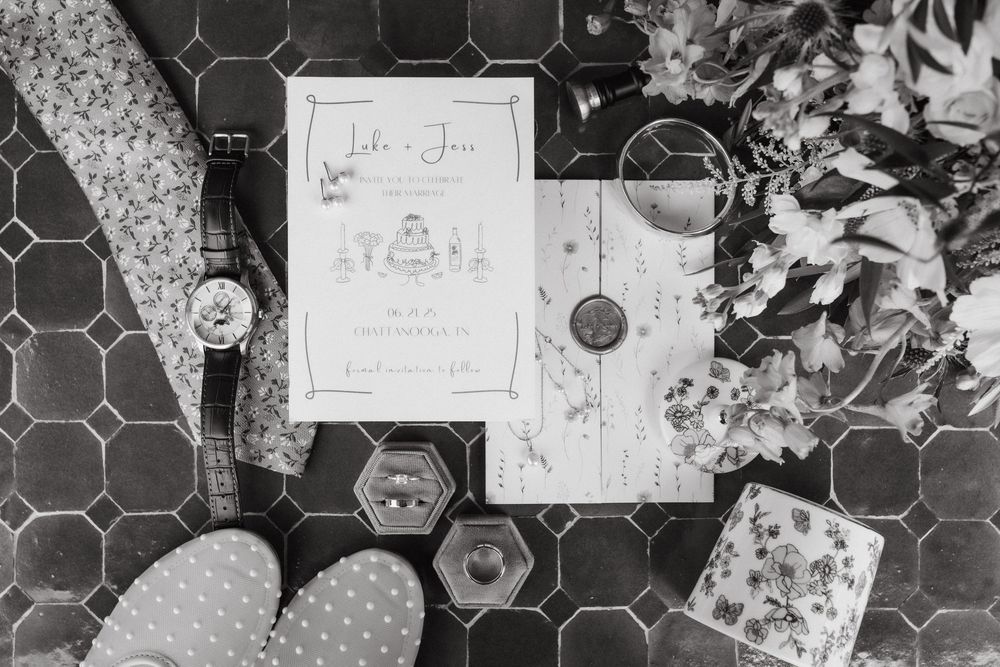 Wedding details flatlay with invitations, rings, and flowers on hexagonal tile in black and white.
