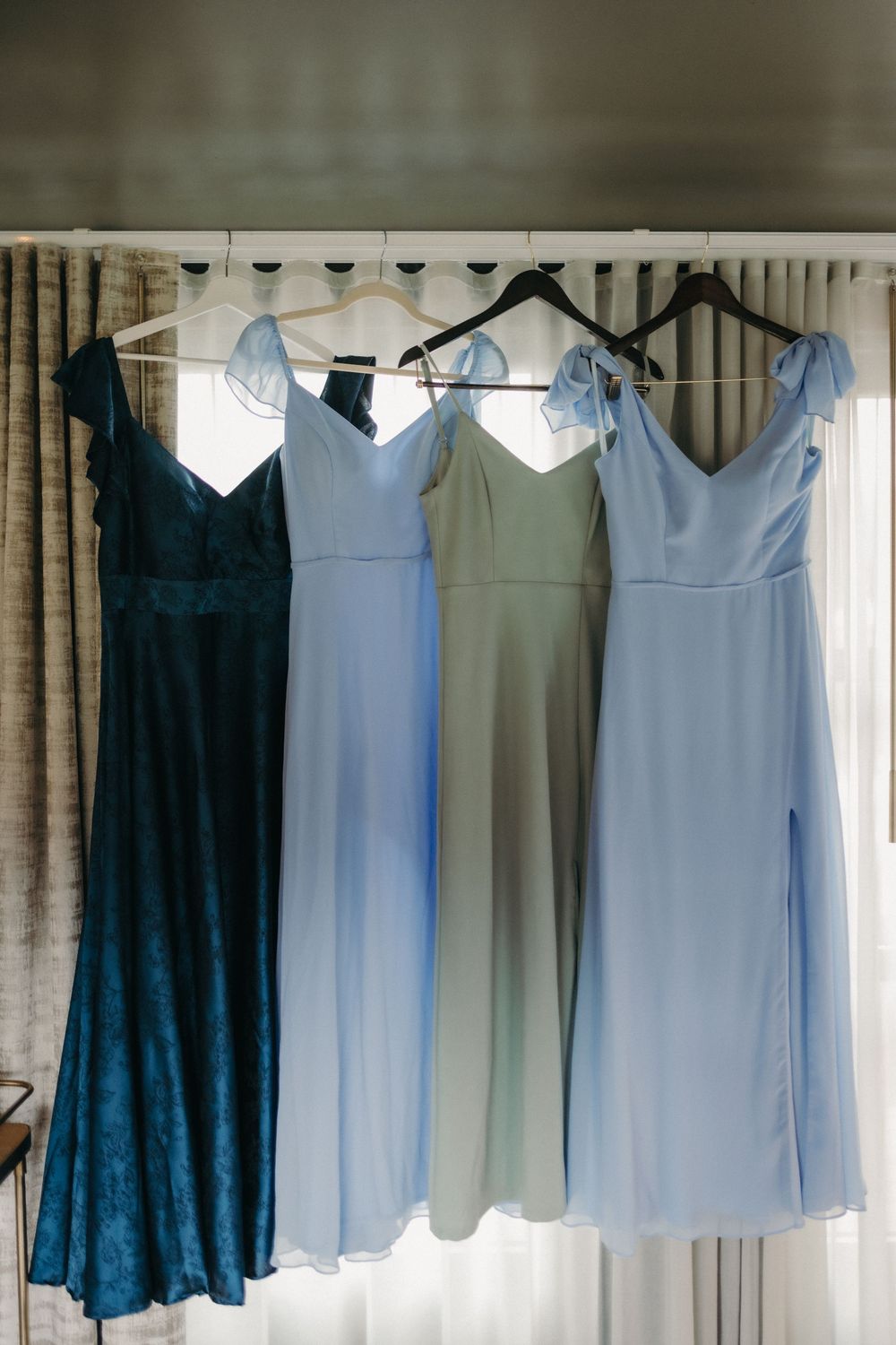 Bridesmaid dresses in shades of blue and navy hanging by a window.