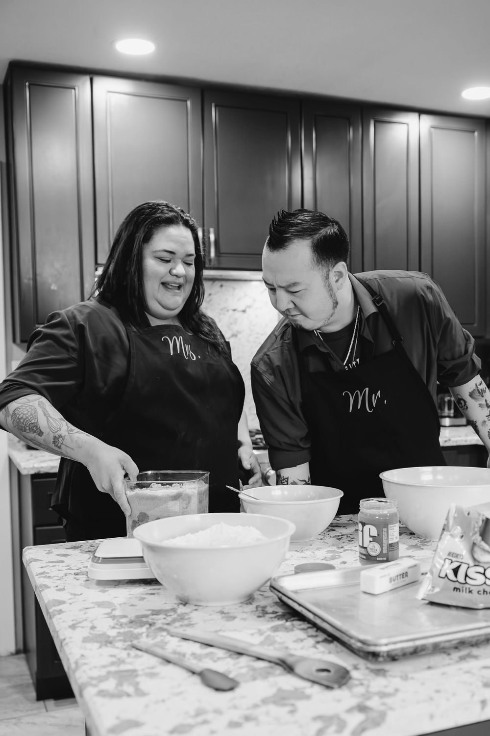 Two people in matching black aprons laugh while cooking together in a modern kitchen with marble countertops.