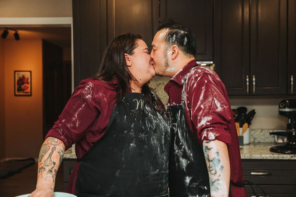 Two people in matching burgundy jackets share a messy chocolate-covered kiss in a kitchen.