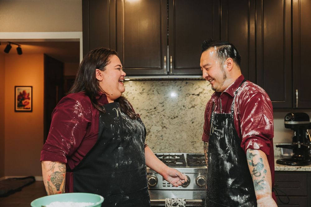 Two people wearing black aprons and burgundy shirts laugh together while cooking in a modern kitchen with dark cabinets.