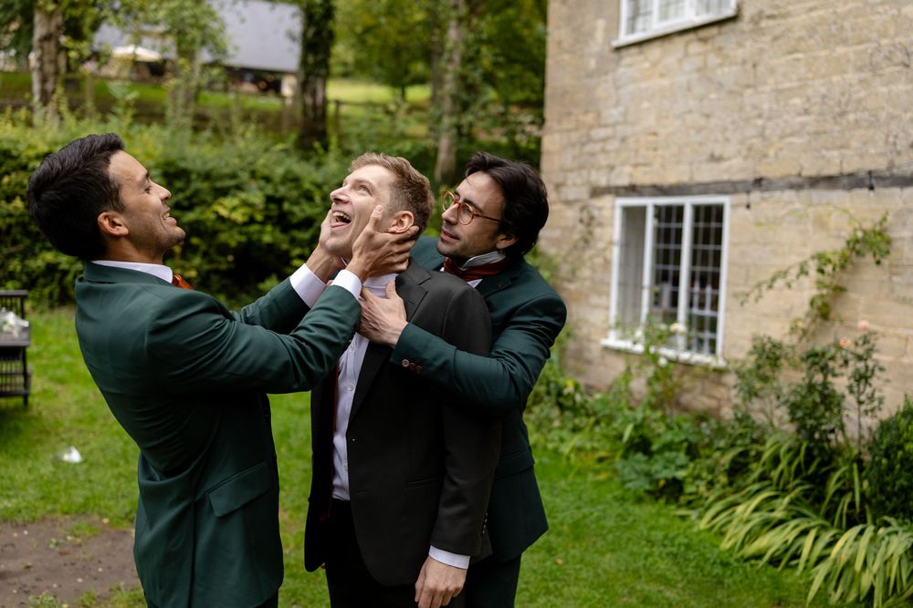 Three friends in suits playfully interact outdoors near a stone building in a garden setting.
