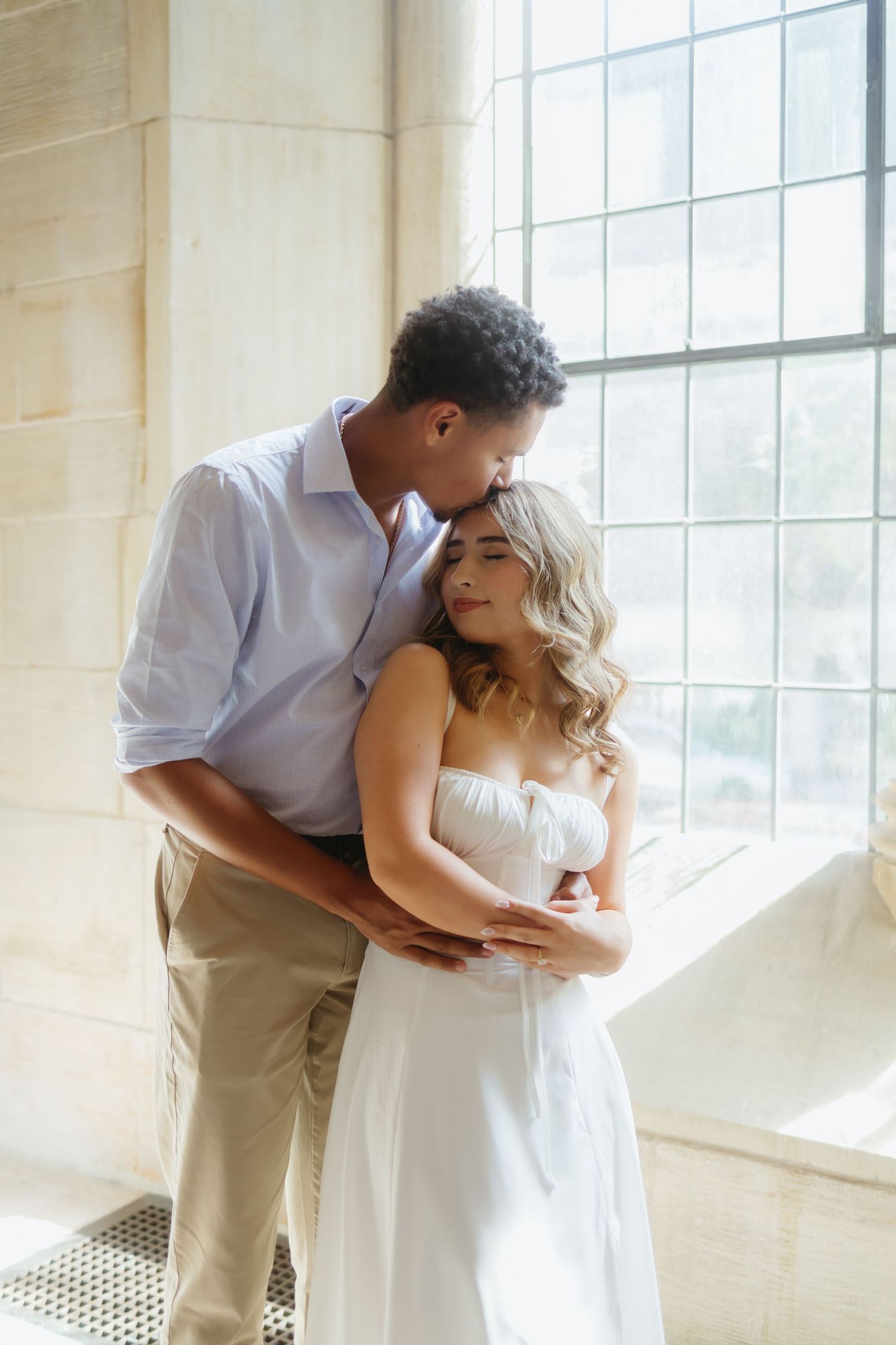 A romantic couple in white clothing embrace by a bright window in a minimalist indoor setting.
