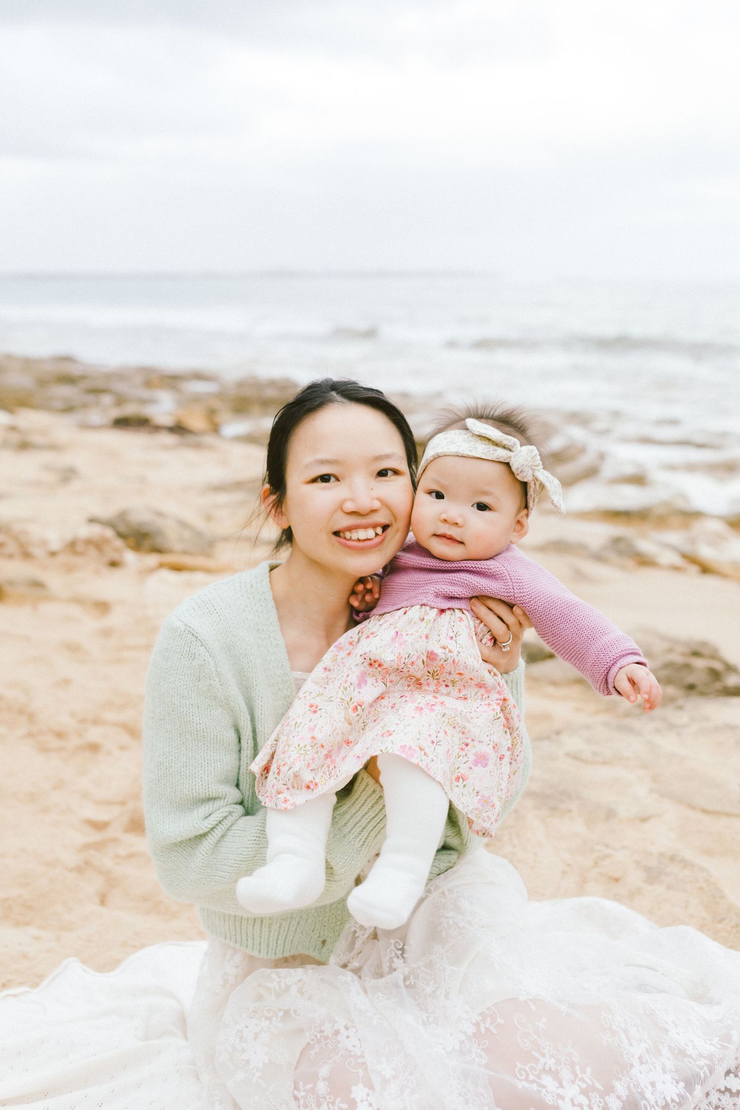 A parent and baby share joyful moments together on a sandy beach with gentle waves in the background.