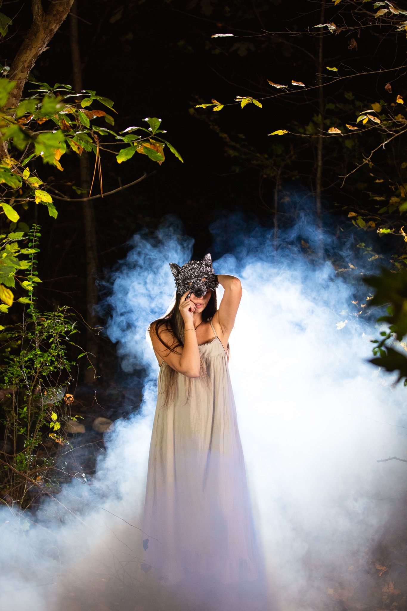 Ethereal scene with white smoke surrounding a woman in a flowing white dress against a dark forest backdrop with a wolf mask.