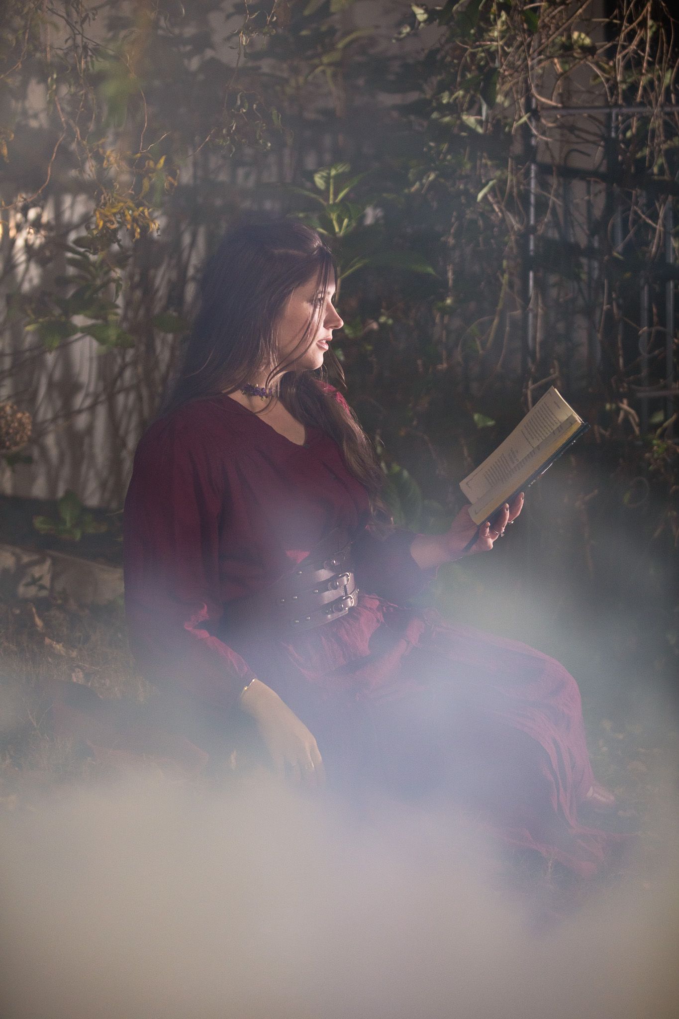 A woman in a red sits outdoors reading a book surrounded by atmospheric fog and greenery.
