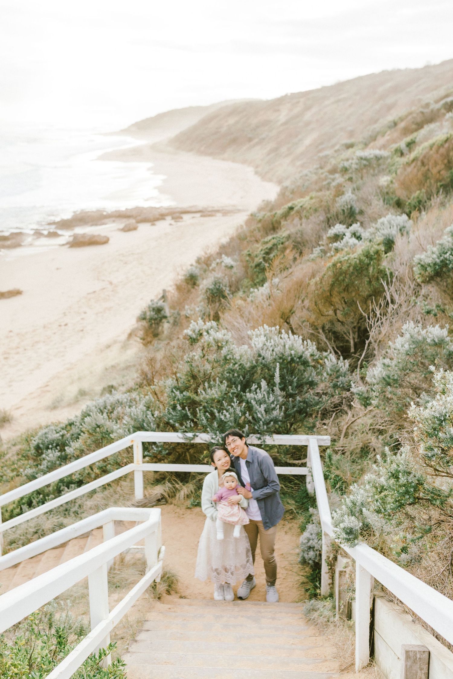 Couple walking along scenic coastal path with white wooden railings and foggy cliffside vegetation in California.