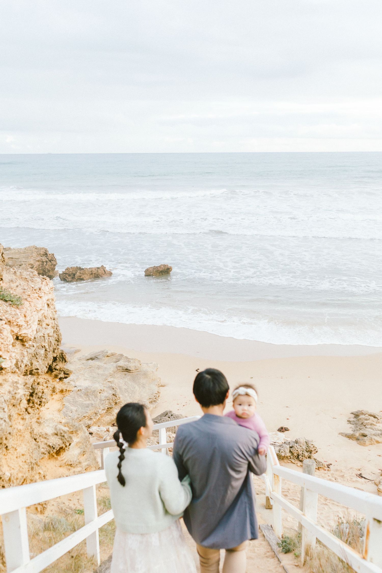 People stand on a white railing overlooking a scenic beach with rocky outcrops and crashing waves on a cloudy day.