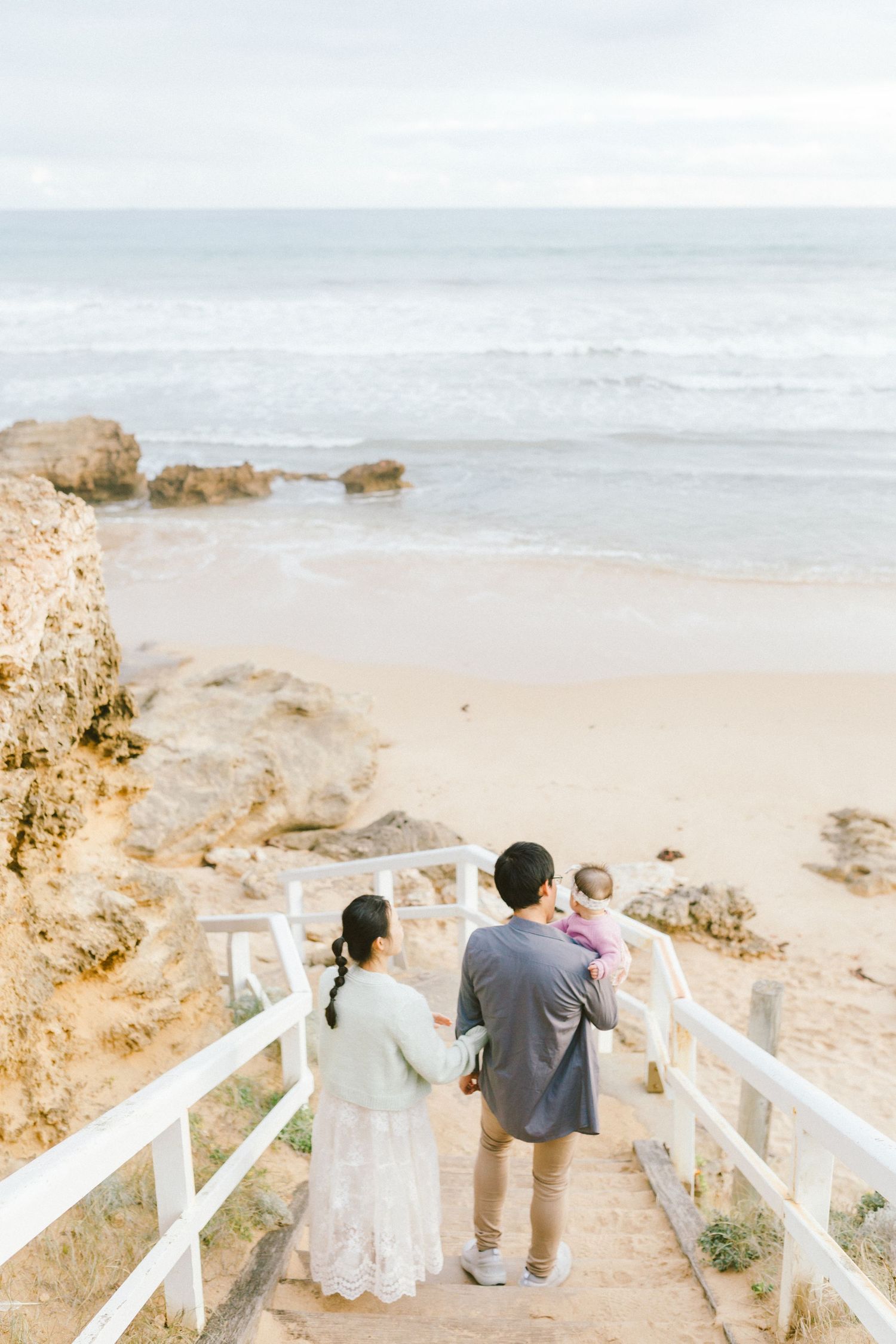 A couple walks down wooden stairs leading to a scenic beach while holding a small child on an overcast day.