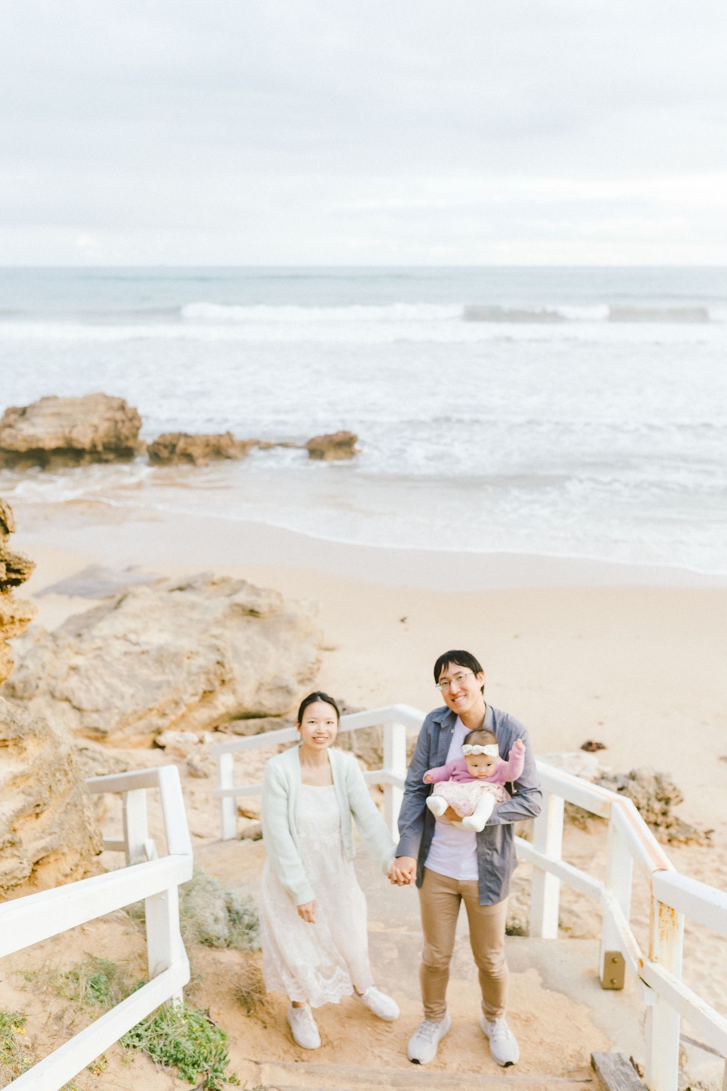 A couple shares a romantic moment along a white fence on a rocky beach with crashing waves in the background.