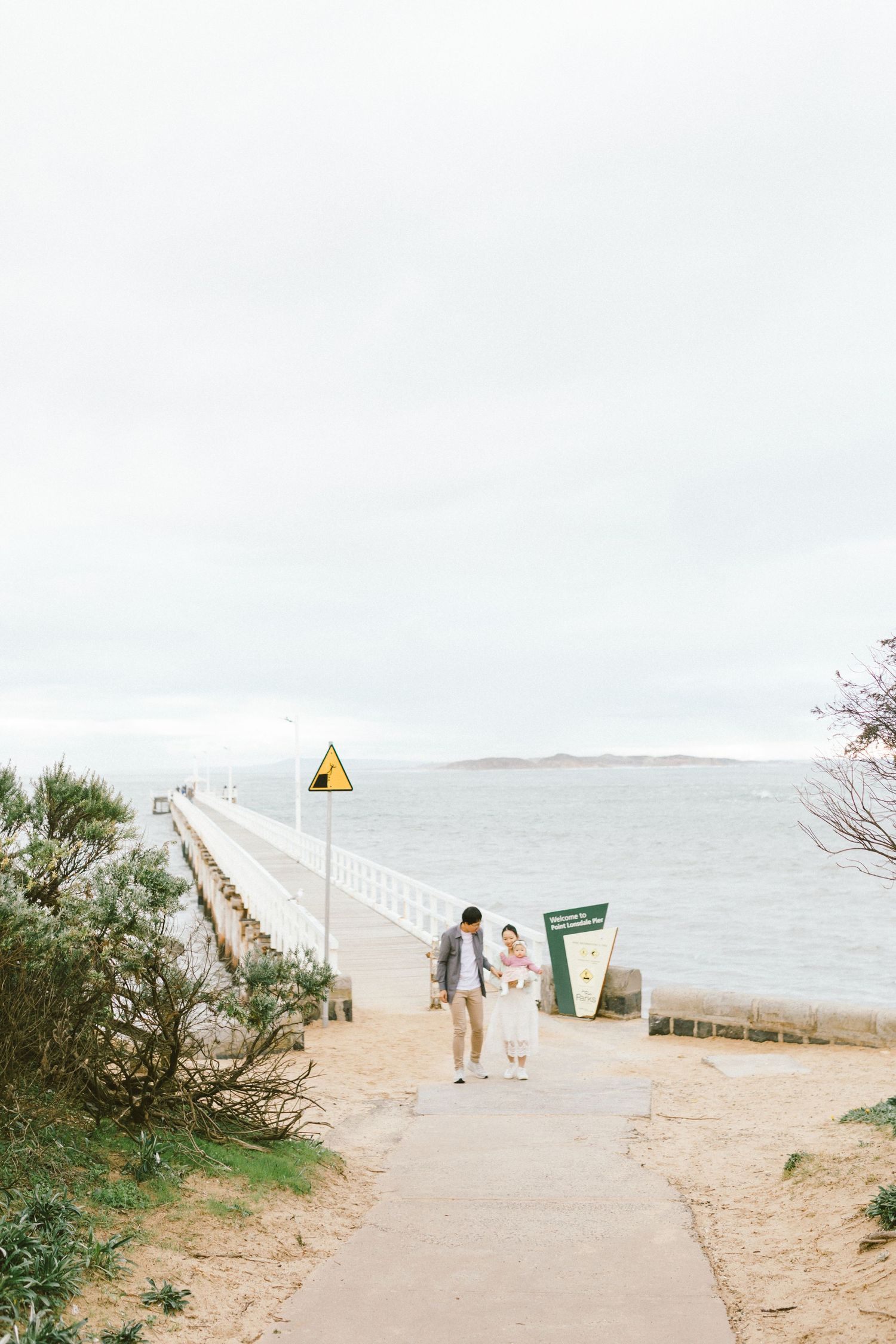 A scenic coastal path leads to a long pier stretching over calm waters on an overcast day.