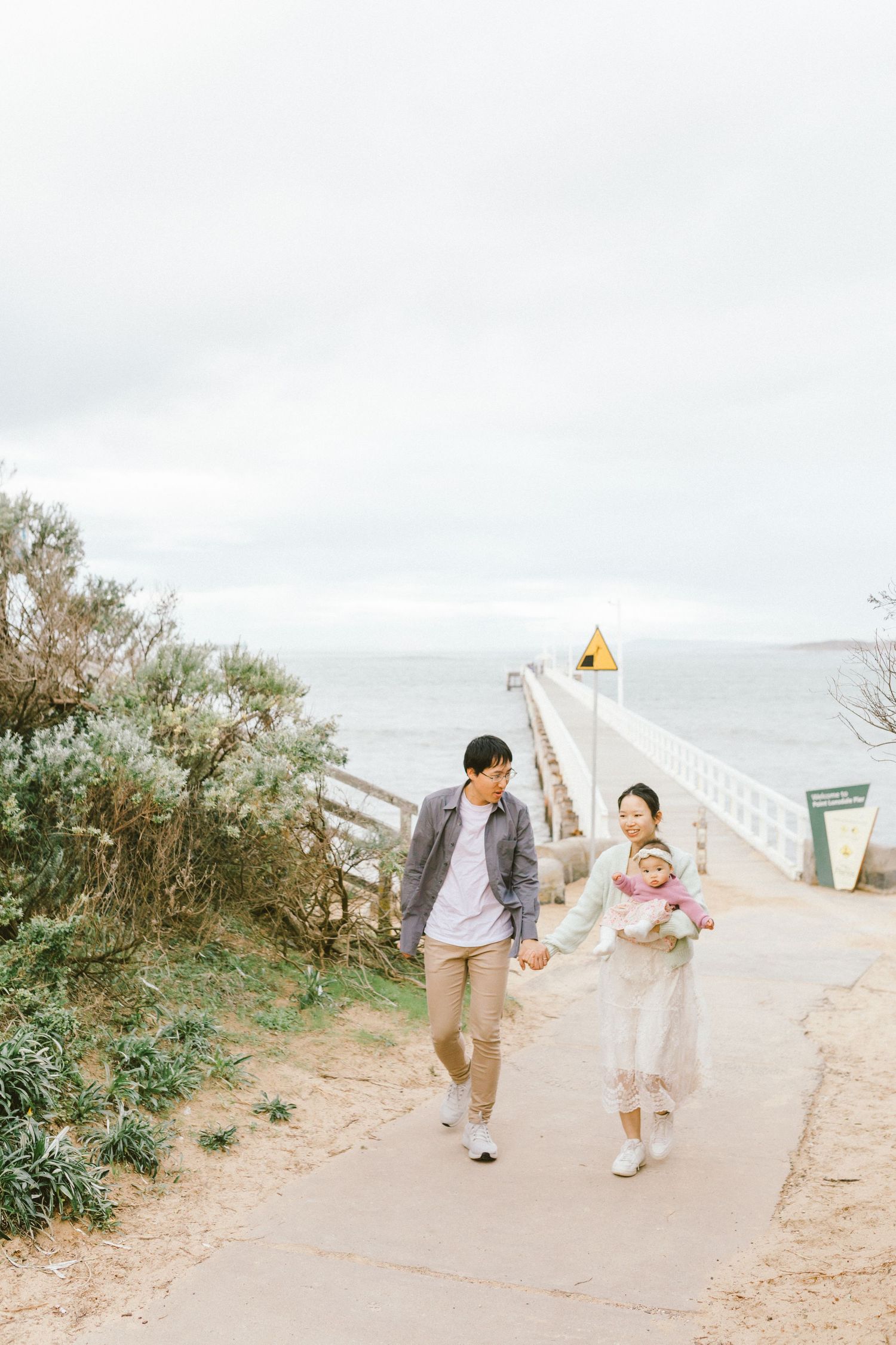People stroll together on a winding beach path surrounded by coastal plants and white fencing.