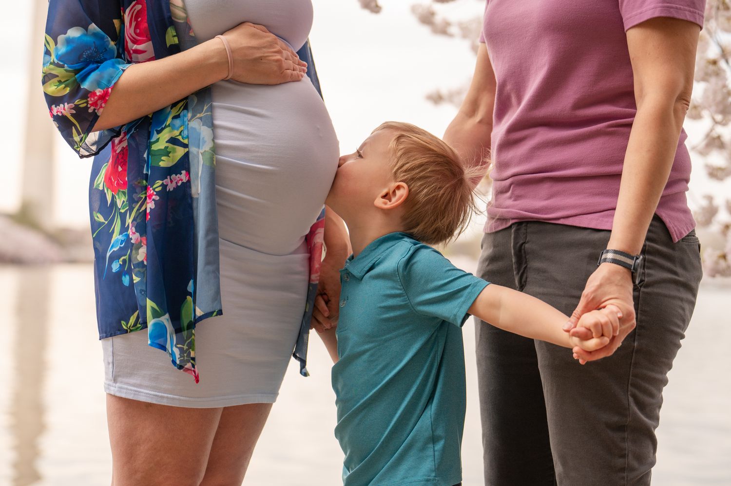 A young boy kisses his pregnant mother's tummy while holding hands with his mom during an outdoor family photo session in Washington DC
