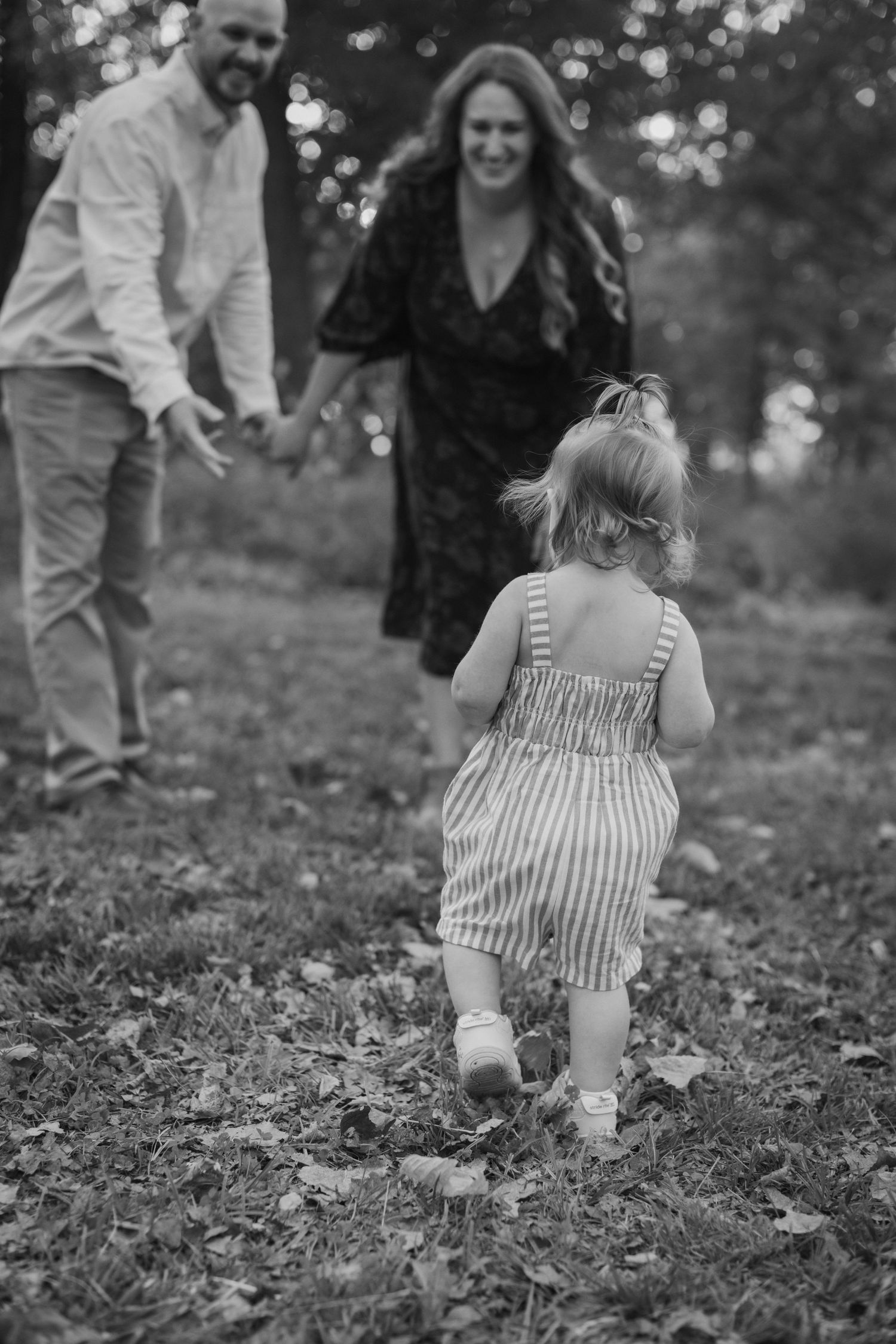 A young toddler runs toward her smiling parents with outstretched arms in a black-and-white image from a Kansas City family photo session.