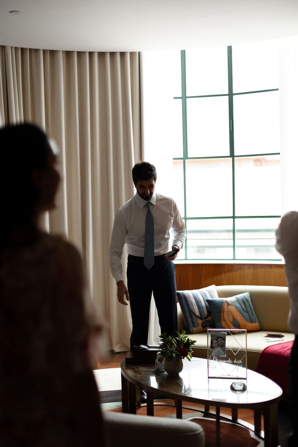 Professional in business attire stands by window in modern office space with couch and coffee table.
