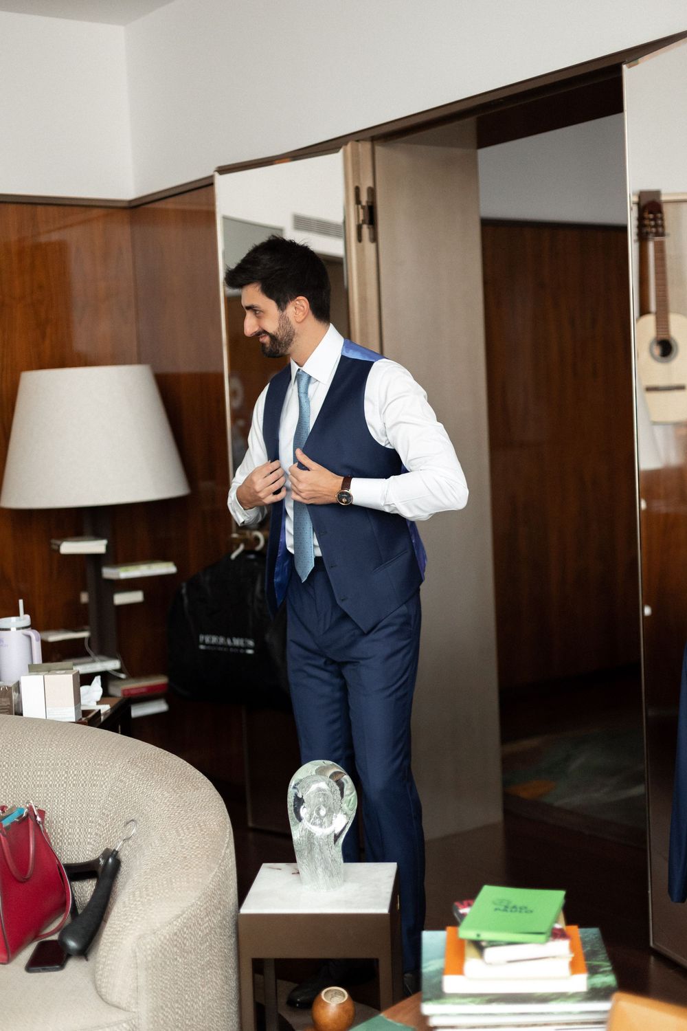 Someone in a navy blue vest and white dress shirt adjusts their attire in a hotel room setting.