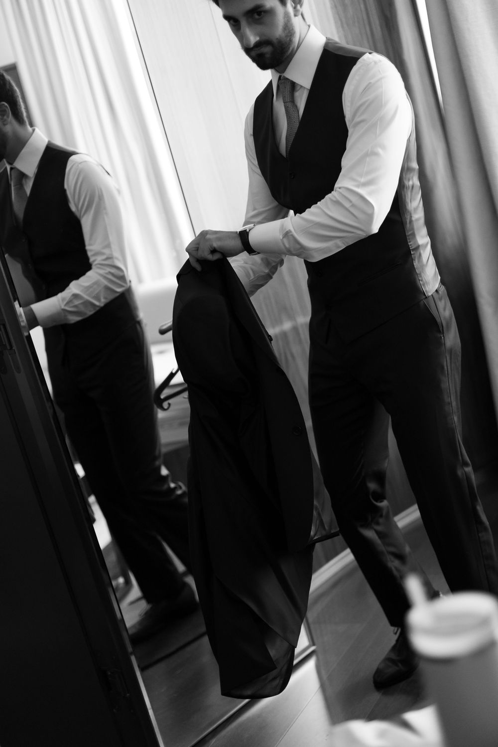 A man in formal attire adjusts his black vest while getting dressed in black and white lighting.
