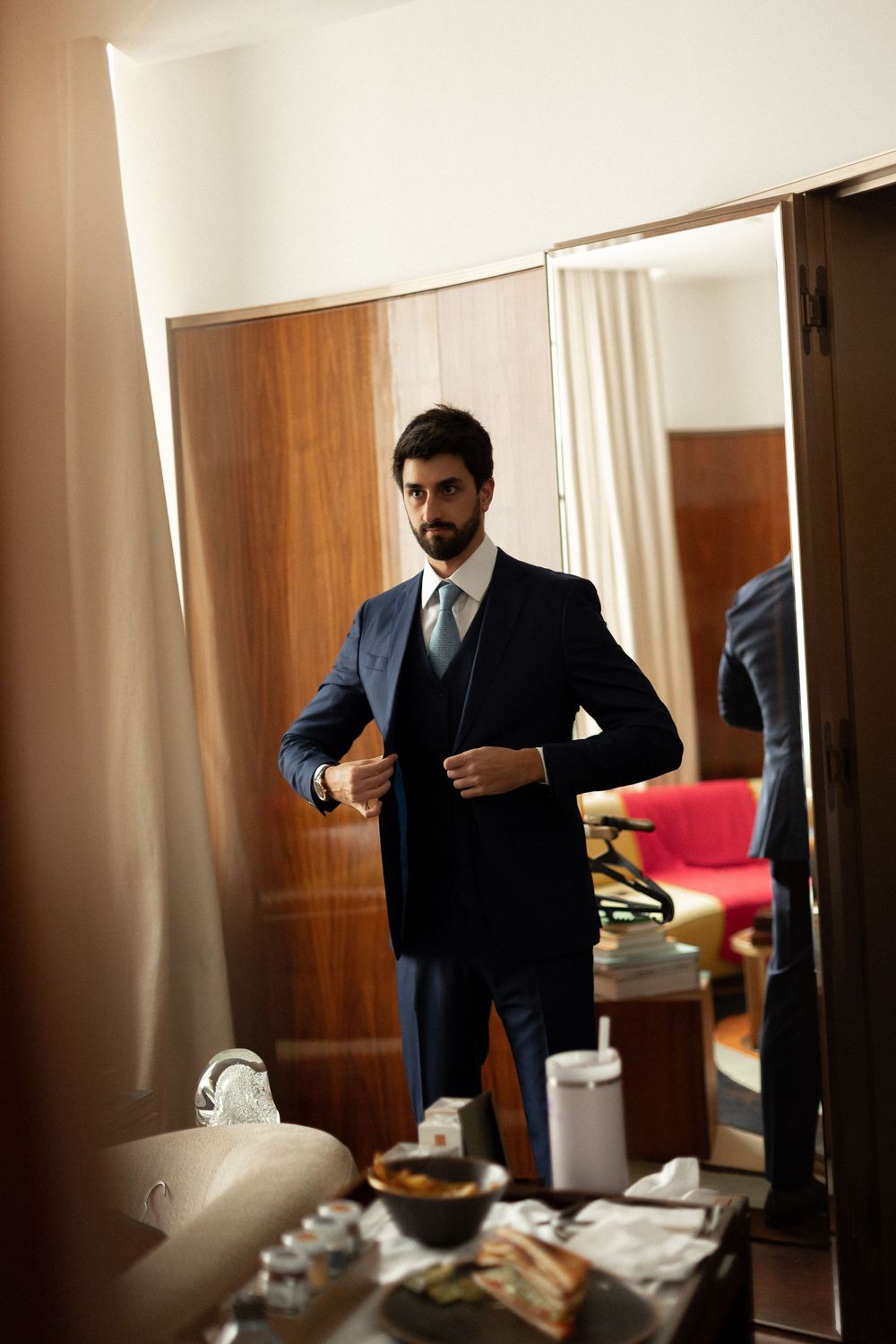 A person in a navy suit adjusts their tie while getting ready in a room with wooden paneling and scattered items.