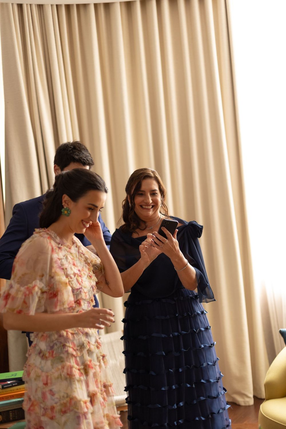 Two people in contrasting dresses share an emotional moment in a room with beige curtains.
