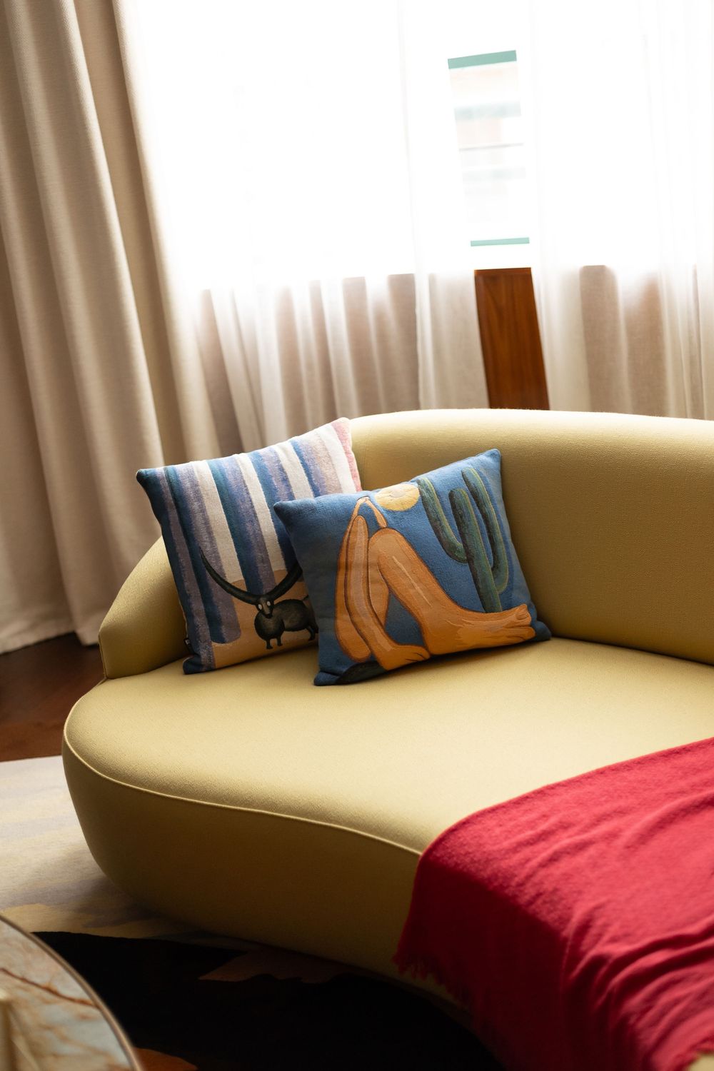 Modern curved sofa with blue throw pillows and red blanket against cream curtains in a contemporary living room.