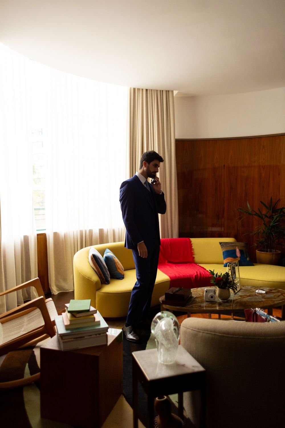 A person in a navy suit stands pensively by a window in a room with yellow and red furniture.