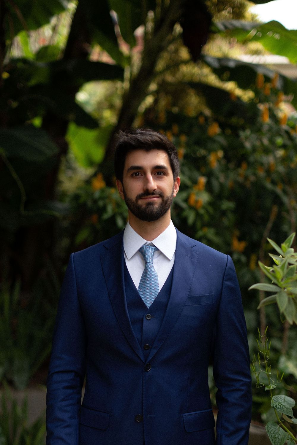 Person in a navy blue three-piece suit poses outdoors against a natural green backdrop.