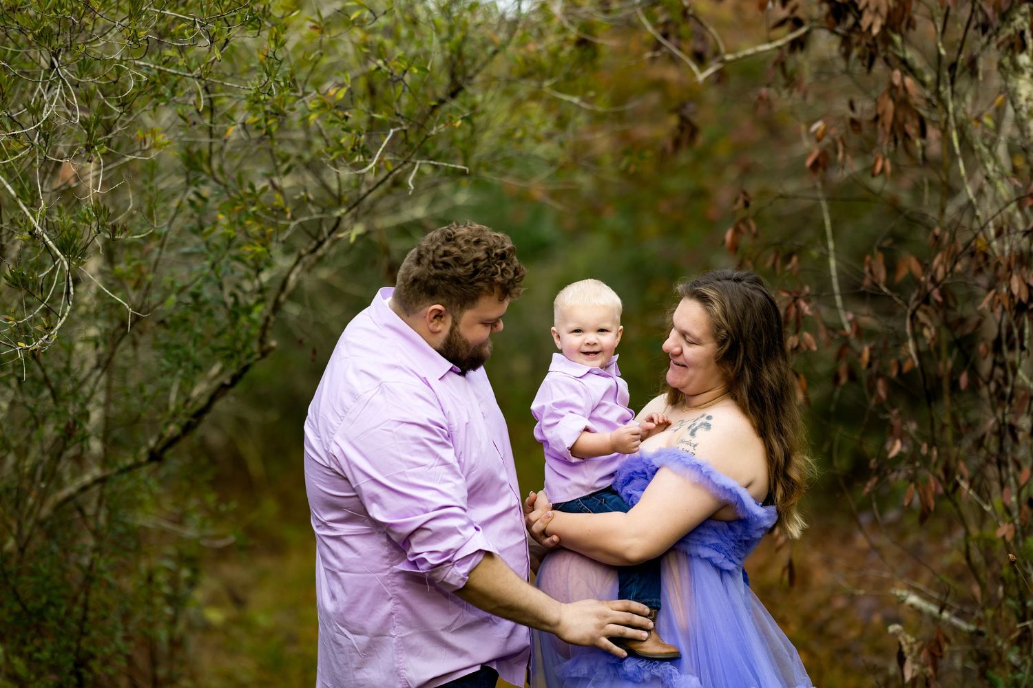 A family in purple and blue outfits poses together in a wooded outdoor setting for professional portraits.