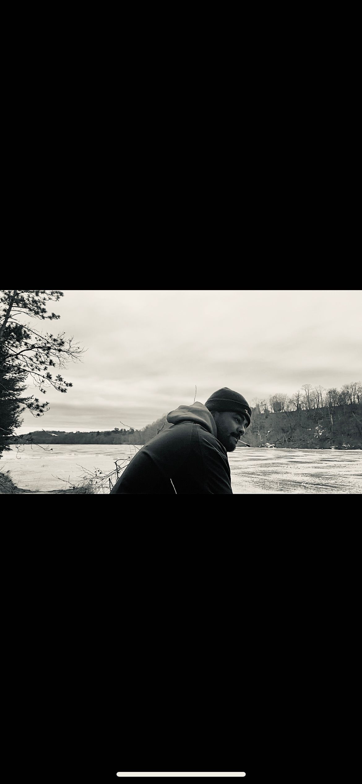 A silhouetted figure crouches near the shoreline of a lake on an overcast day with trees in the background.