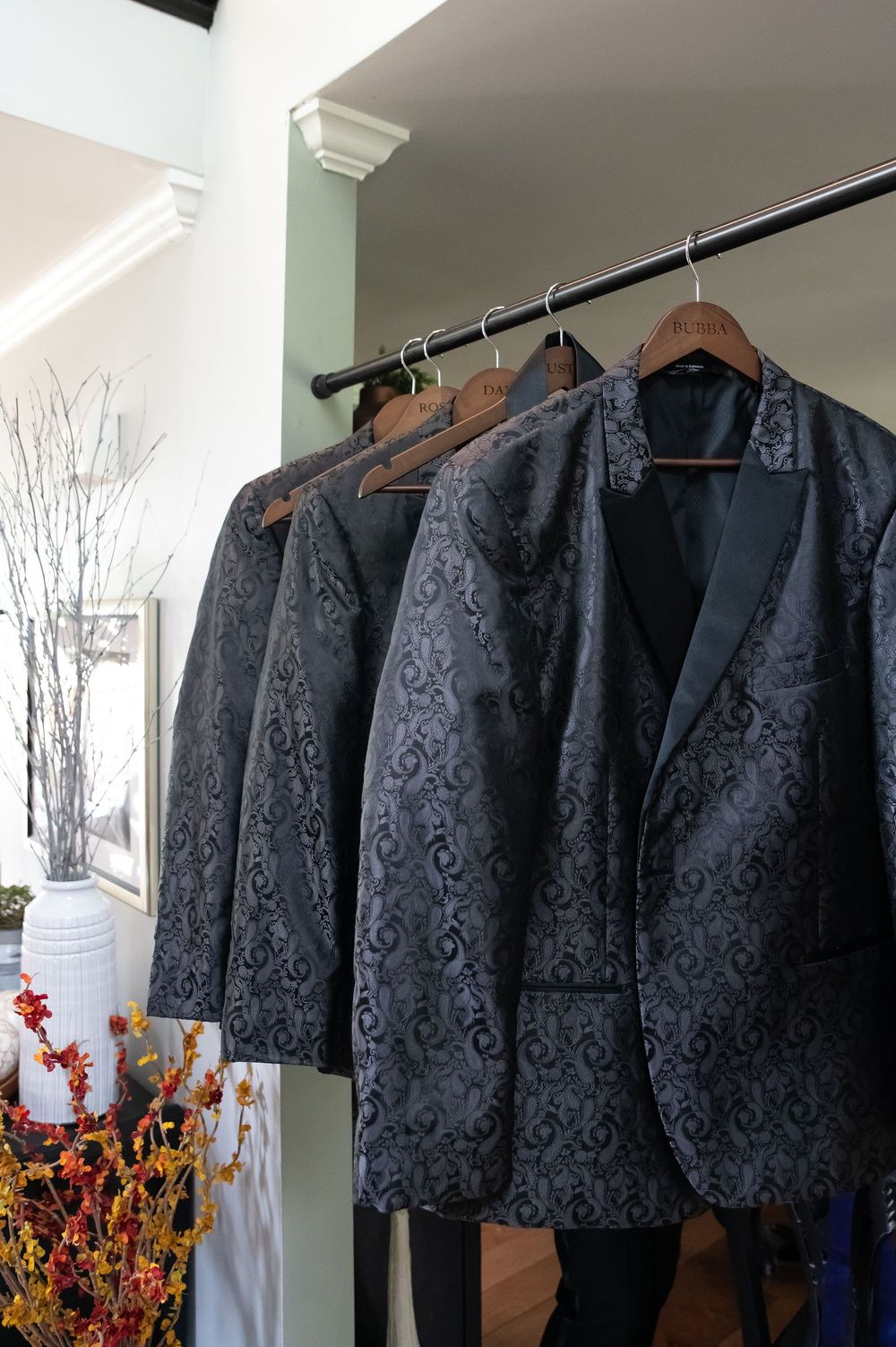 Black damask jackets hanging on wooden hangers in a closet with autumn floral decorations visible.