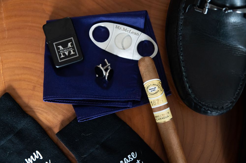 Luxury cigar and cutter displayed with navy blue accessories and black leather loafer on wooden surface.