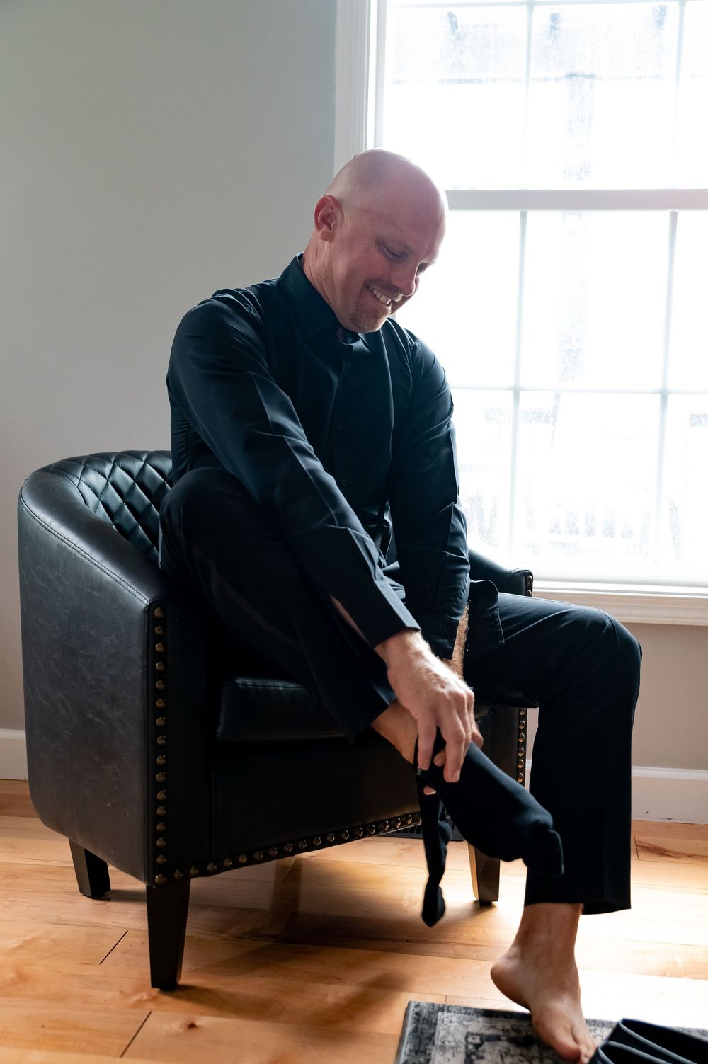 Someone wearing all black sits casually in a quilted leather accent chair near a bright window on hardwood floors.