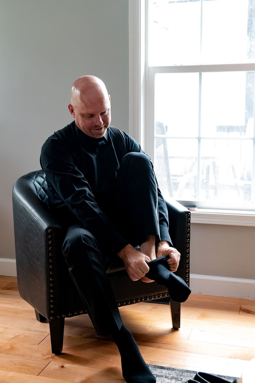 Man in black clothing sits on dark leather chair in bright room with hardwood floors and sunlit windows.