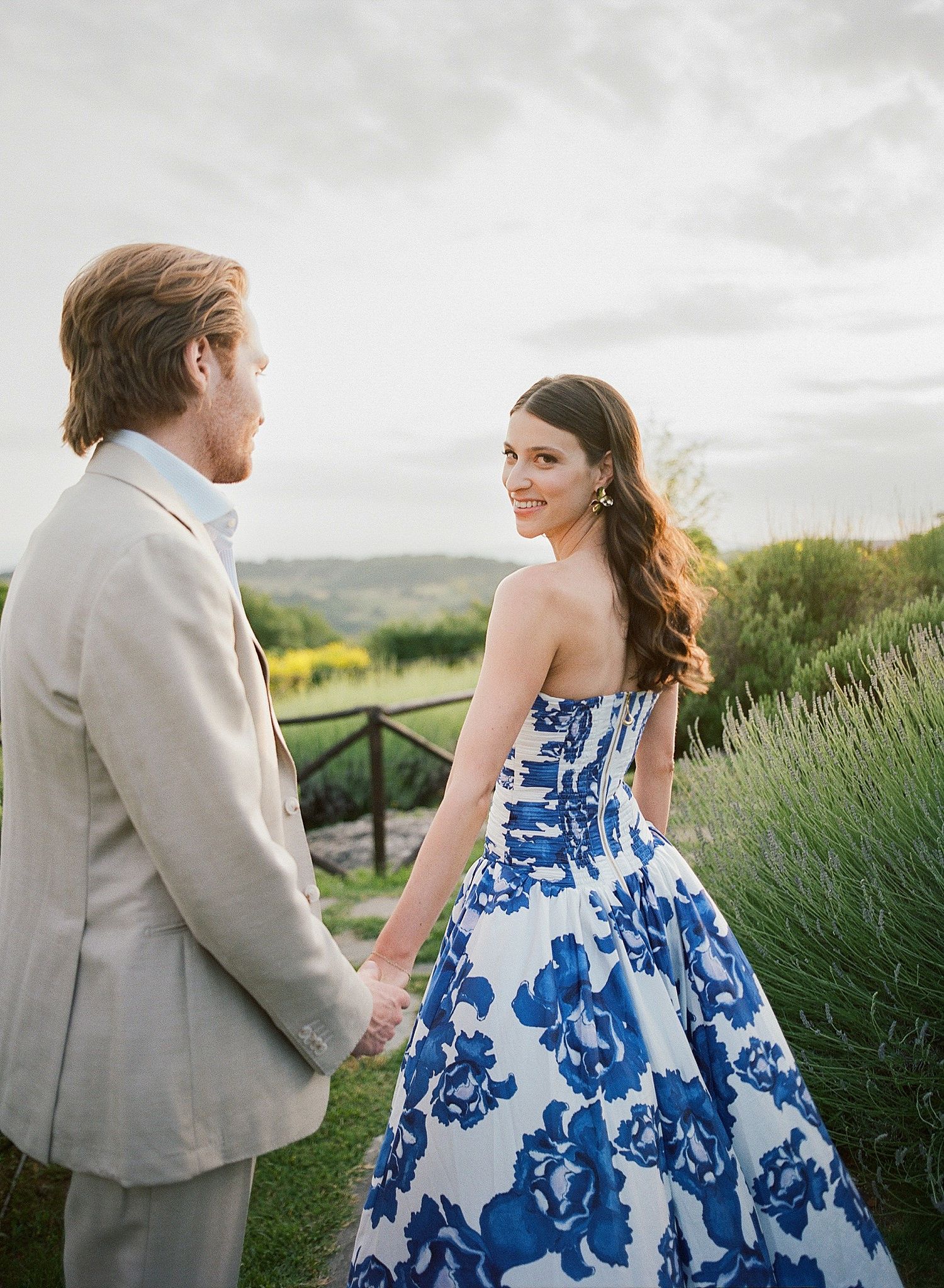 A couple in wedding attire pose in a garden setting with the bride wearing a stunning blue and white floral gown.