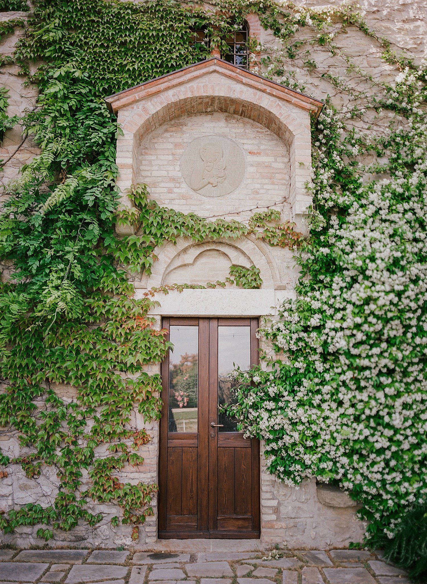Rustic wooden door with stone archway surrounded by lush green ivy and white flowering vines on old stone wall.