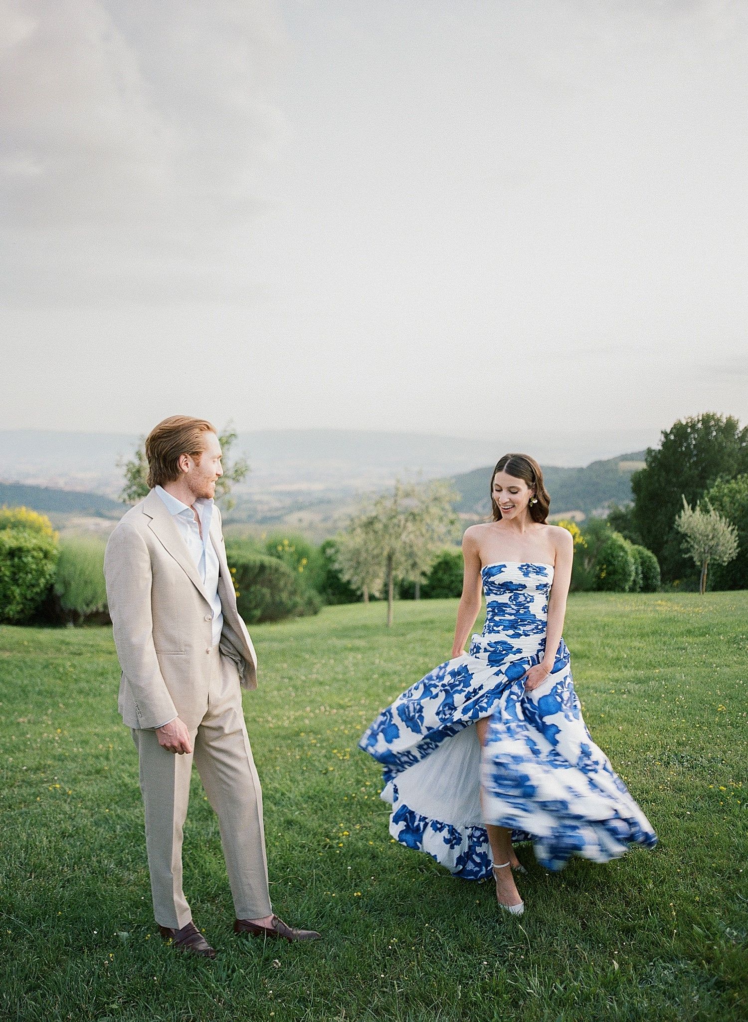 A couple dances playfully in a grassy hilltop meadow, with the bride wearing a blue and white floral dress.