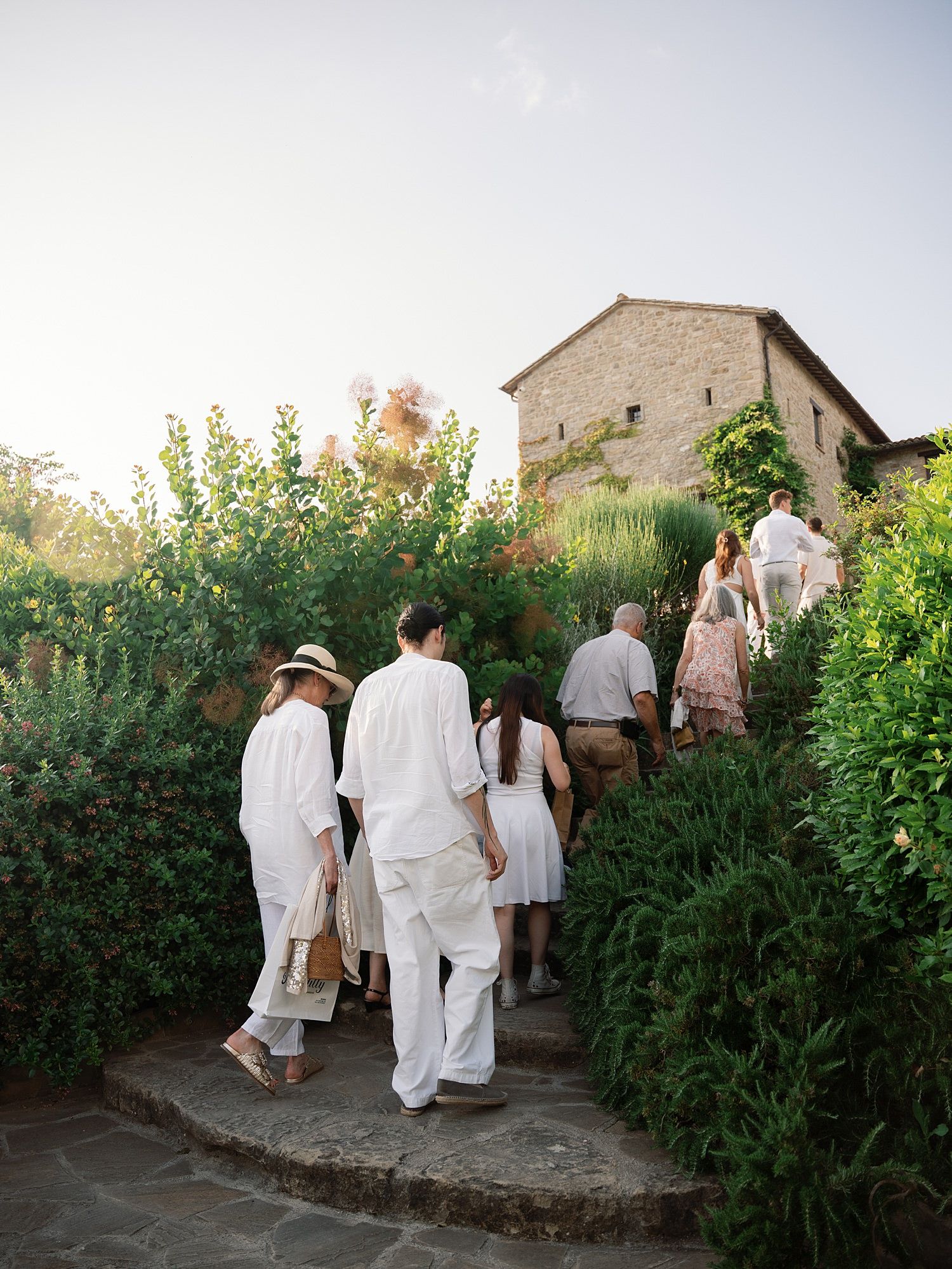 People in white clothing walking along a garden path beside stone buildings in a rustic setting.