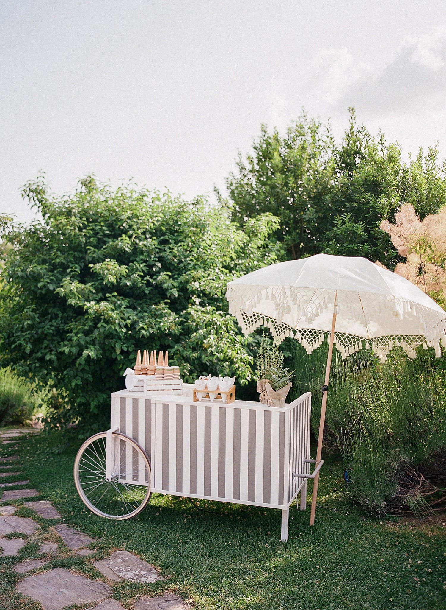 Elegant outdoor party setup with white parasols, vintage bicycle, and striped tablecloths in a lush garden setting.