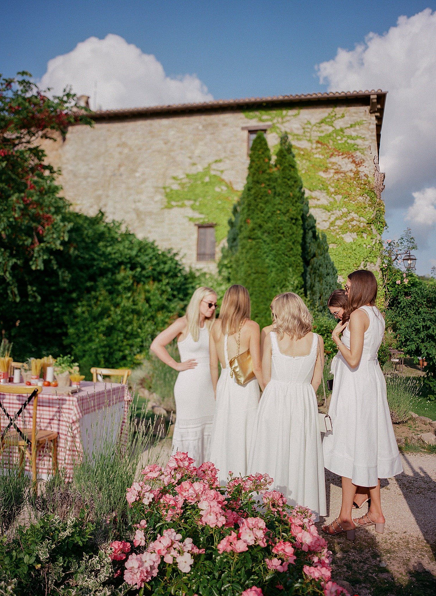 Four people in white dresses gather in a garden with pink roses and lavender near a stone wall on a sunny day.