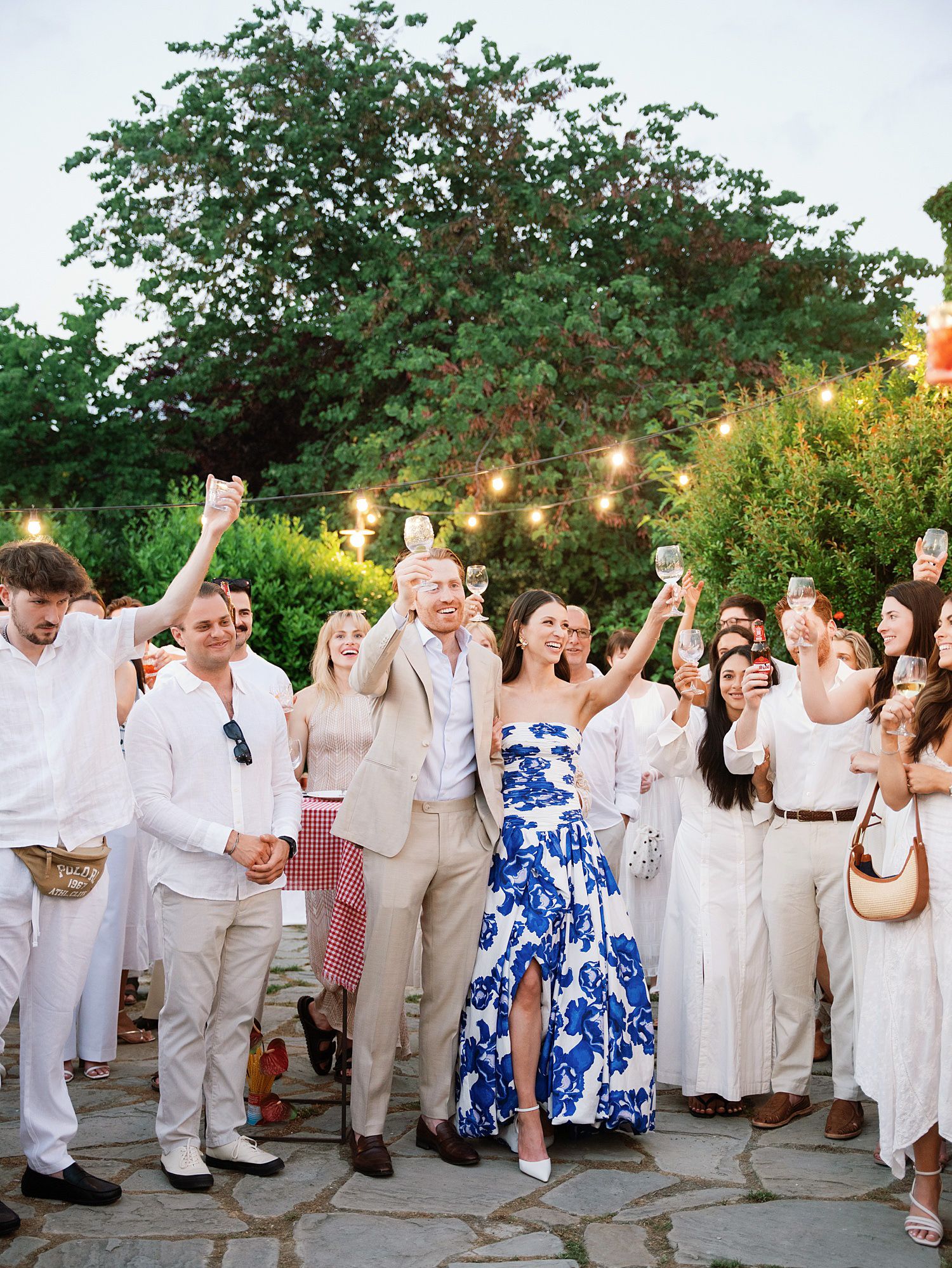 Wedding guests in white and blue attire celebrate with raised glasses during an outdoor reception at sunset.